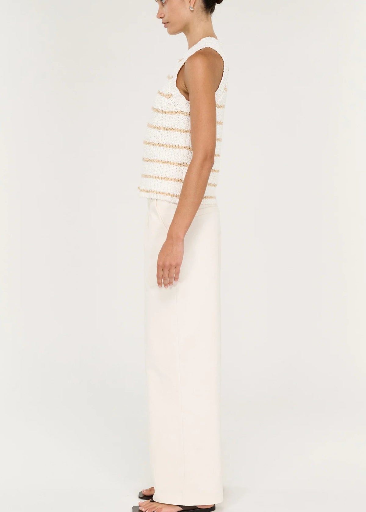 A model stands against a plain light background wearing a sleeveless knit top with thin horizontal beige stripes on an off-white base, paired with off-white wide-leg pants and black sandals.