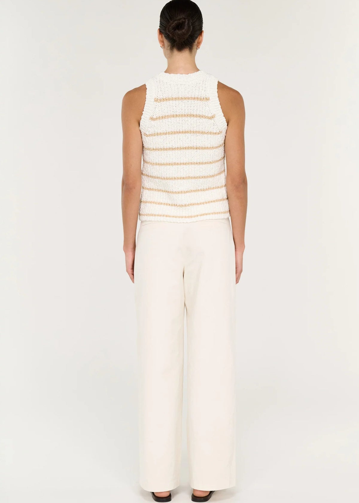 A model stands against a plain light background wearing a sleeveless knit top with thin horizontal beige stripes on an off-white base, paired with off-white wide-leg pants and black sandals.