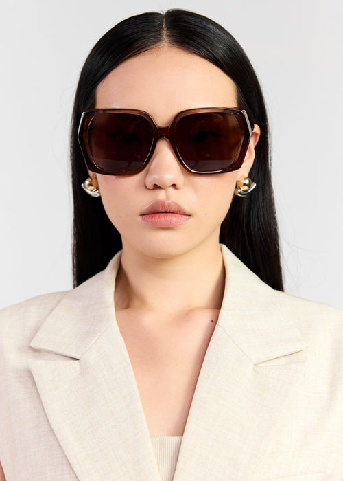 A model with long black hair is wearing oversized square sunglasses with dark brown frames and dark brown lenses, paired with a light beige blazer. She is photographed against a clean, light gray background.