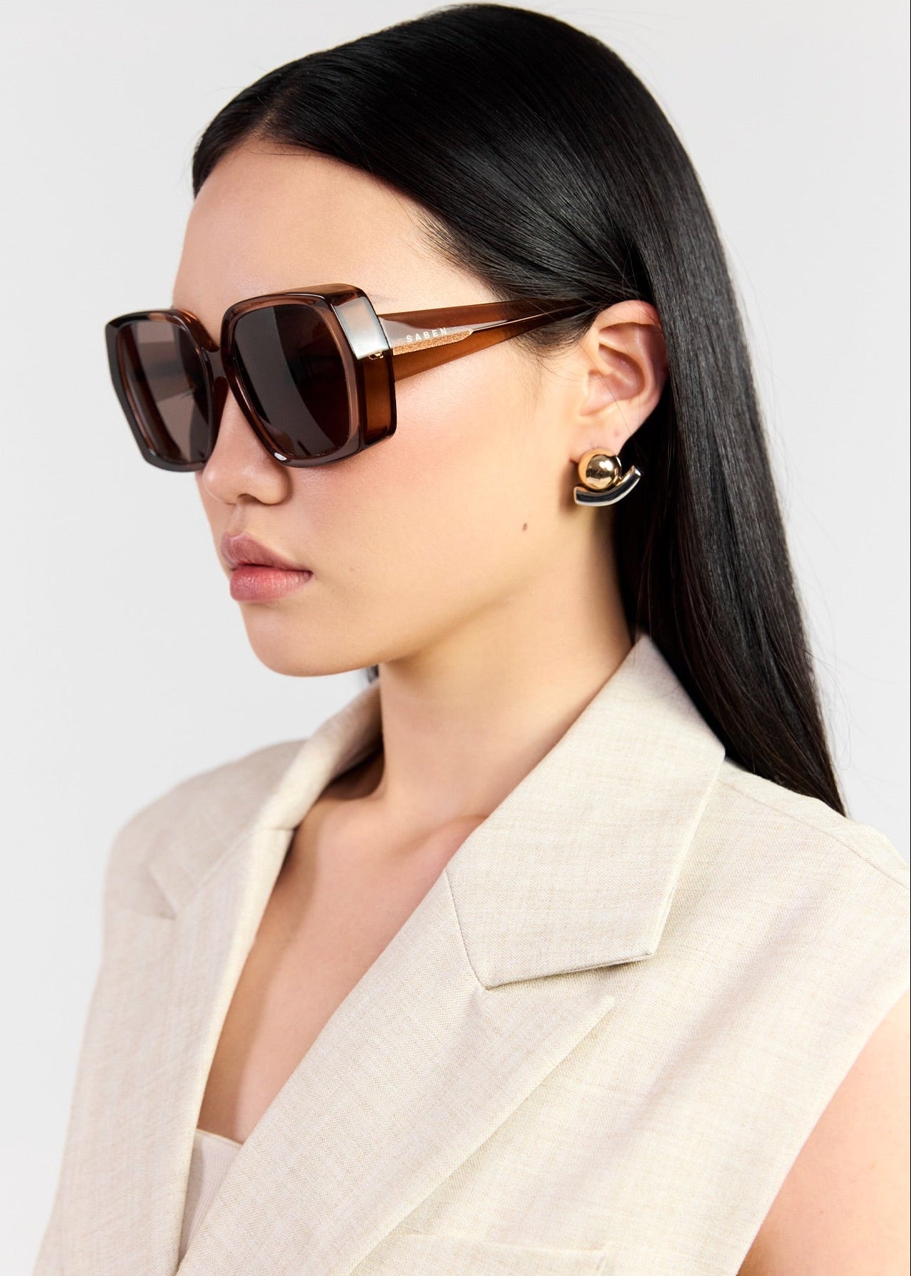 A model with long black hair is wearing oversized square sunglasses with dark brown frames and dark brown lenses, paired with a light beige blazer. She is photographed against a clean, light grey background.