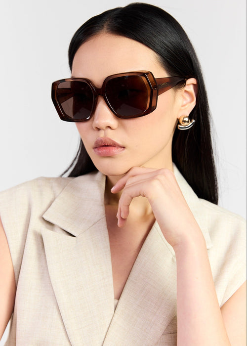 A model with long black hair is wearing oversized square sunglasses with dark brown frames and dark brown lenses, paired with a light beige blazer. She is photographed against a clean, light grey background.
