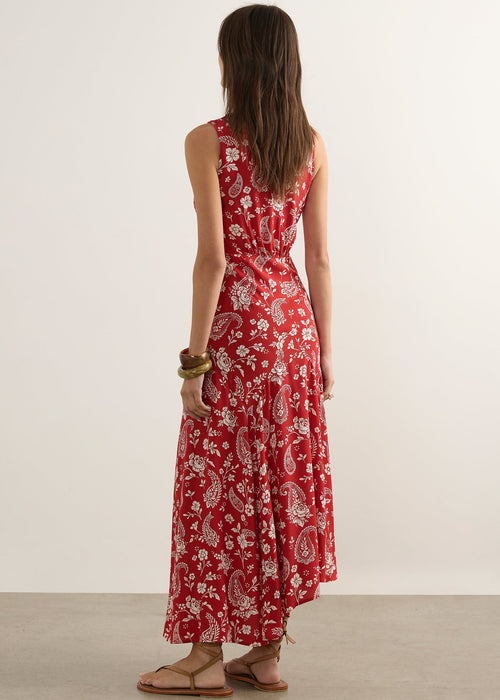 A woman stands with her back to the camera wearing a sleeveless red maxi dress with a white paisley and floral pattern, paired with brown sandals, in a minimalist studio setting.