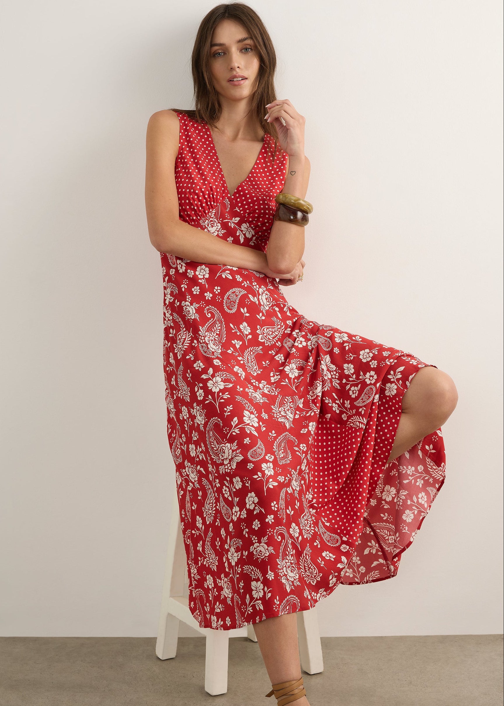 A woman modelling a sleeveless red maxi dress with a white paisley and floral print, standing against a plain off-white studio wall.