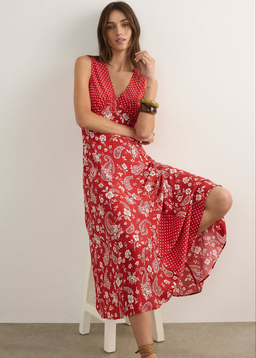 A woman modelling a sleeveless red maxi dress with a white paisley and floral print, standing against a plain off-white studio wall.
