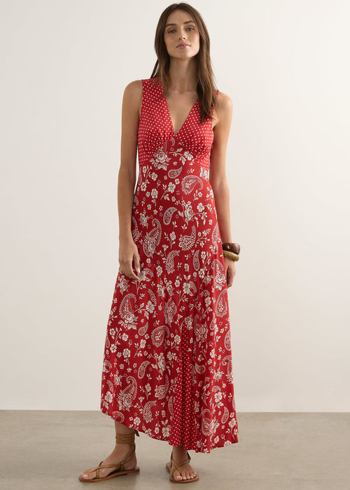 A woman modelling a sleeveless red maxi dress with a white paisley and floral print, standing against a plain off-white studio wall.