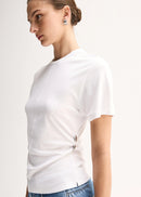 A woman wearing a white short-sleeve t-shirt with a unique wrap-style hem and small metallic button details, paired with blue jeans, photographed against a clean off-white background.
