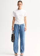 A model stands against a plain light background wearing a white short-sleeve top, wide-leg blue jeans, and holding a dark brown leather handbag.