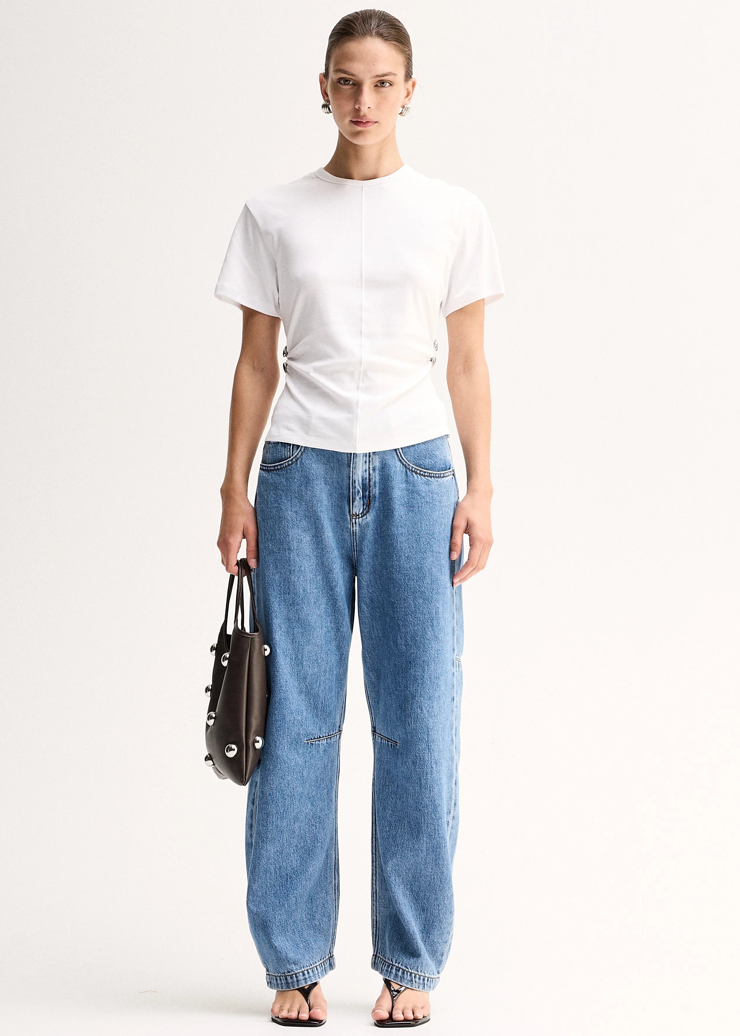 A model stands against a plain light background wearing a white short-sleeve top, wide-leg blue jeans, and holding a dark brown leather handbag.
