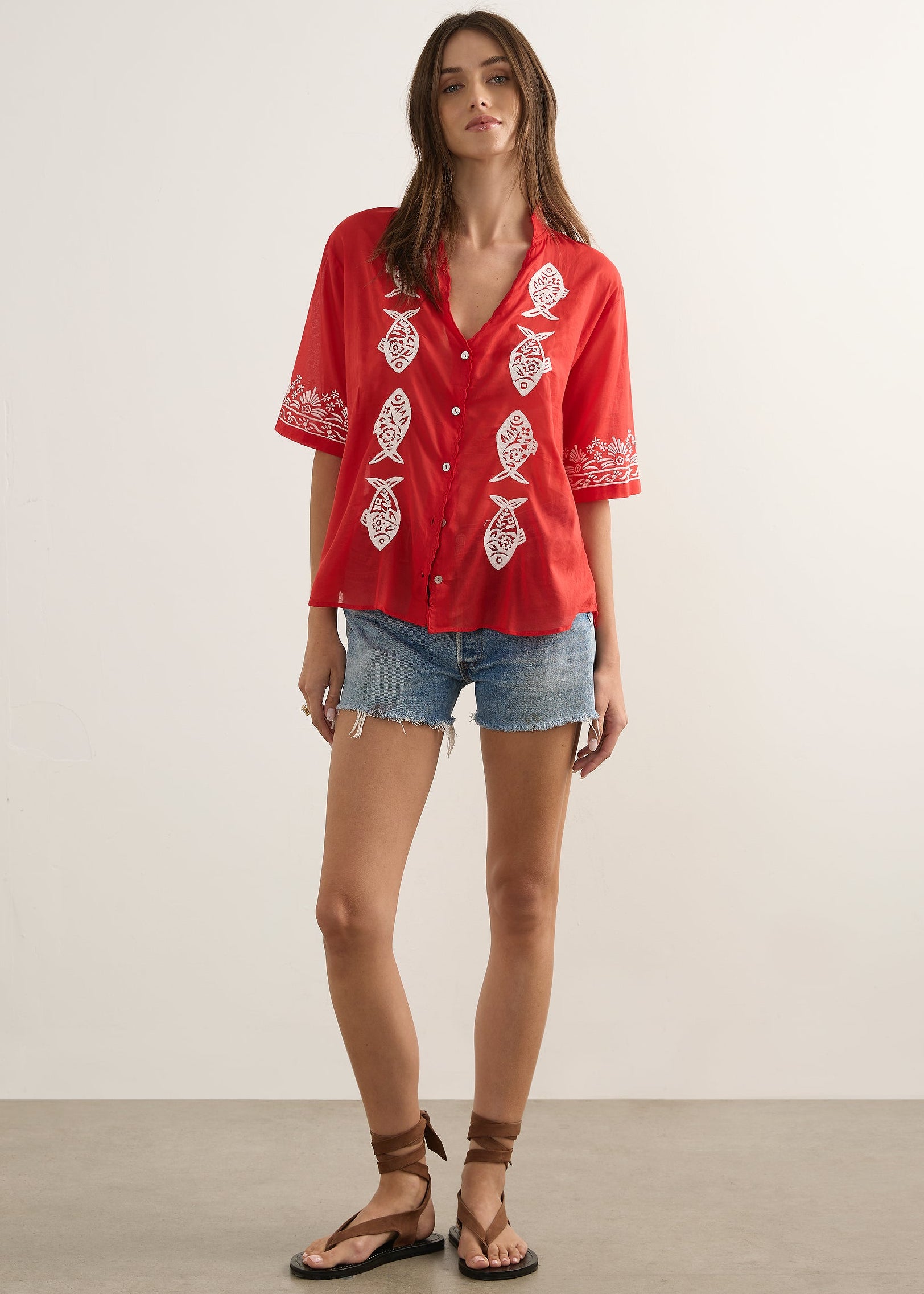 A woman modeling a red short-sleeve button-up shirt with white embroidered fish motifs, paired with denim shorts and brown sandals, standing against a plain light beige studio background.