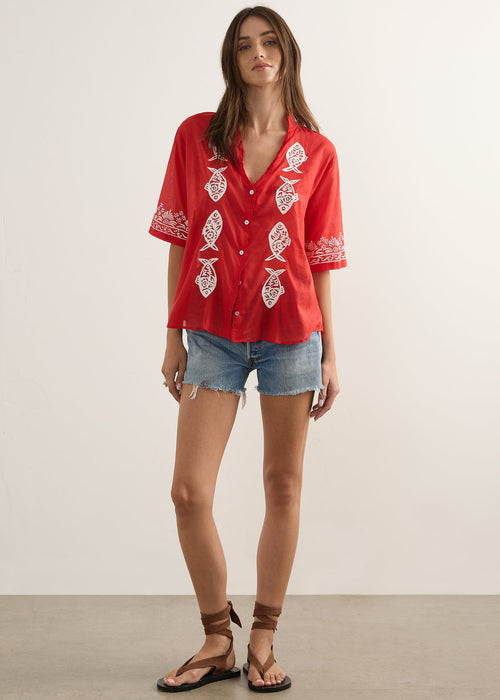 A woman modeling a red short-sleeve button-up shirt with white embroidered fish motifs, paired with denim shorts and brown sandals, standing against a plain light beige studio background.