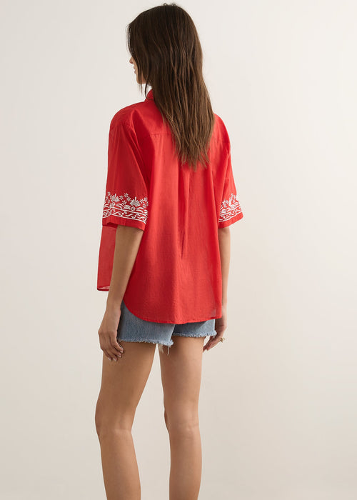 A woman modelling a red short-sleeve button-up shirt with white embroidered fish motifs, paired with denim shorts and brown sandals, standing against a plain light beige studio background.