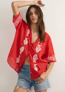 A woman modelling a red short-sleeve button-up shirt with white embroidered fish motifs, paired with denim shorts and brown sandals, standing against a plain light beige studio background.