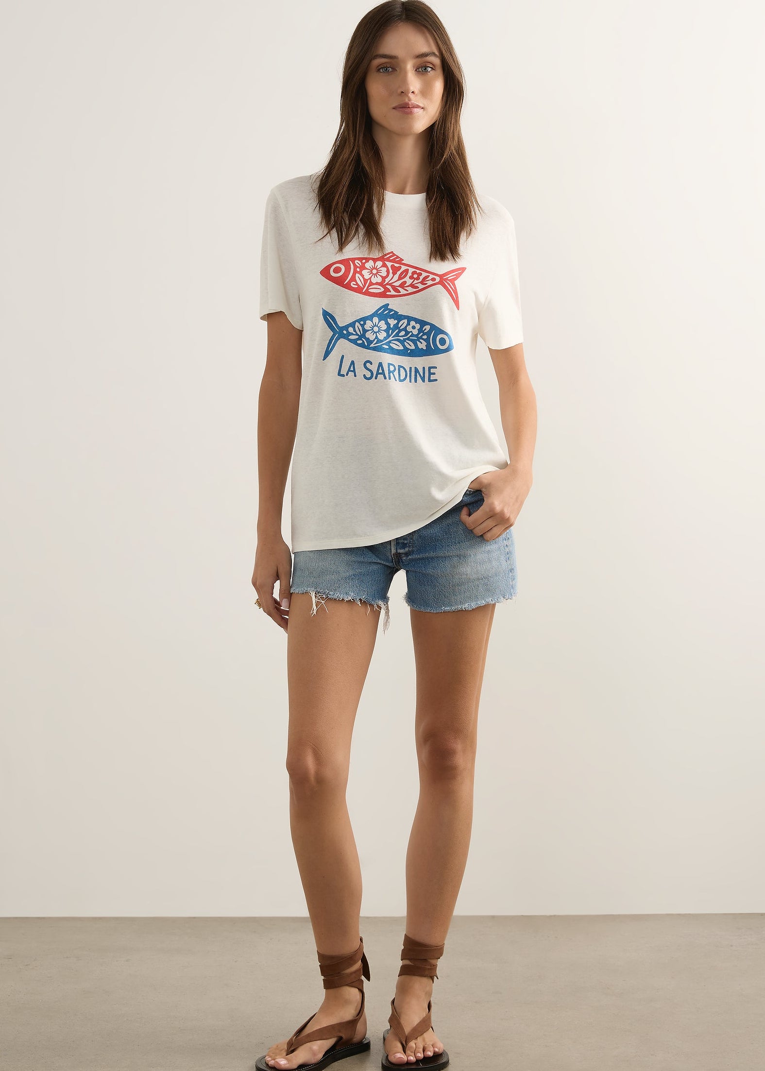 A woman modelling a white graphic t-shirt with a red and blue fish design and the text 'LA SAINE' and 'CARNICERIE', paired with denim shorts and brown sandals, standing against a plain light grey studio background.
