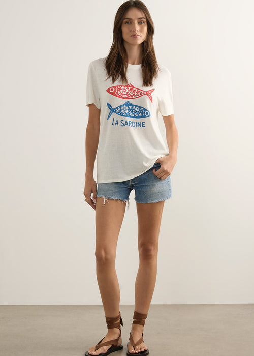 A woman modelling a white graphic t-shirt with a red and blue fish design and the text 'LA SAINE' and 'CARNICERIE', paired with denim shorts and brown sandals, standing against a plain light grey studio background.