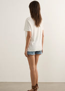 A woman is shown from the back wearing a white short-sleeve t-shirt, denim shorts, and brown strappy sandals in a minimalist studio setting with a warm off-white wall and light grey floor.