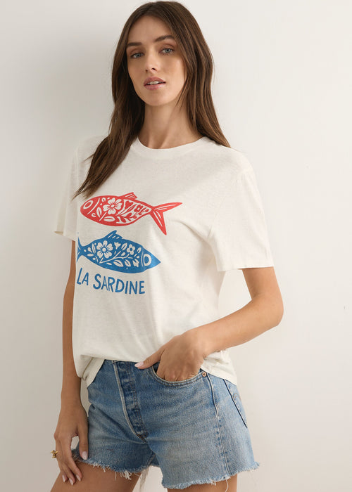 A woman modelling a white graphic t-shirt with a red and blue fish design and the text 'LA SAINE' and 'CARNICERIE', paired with denim shorts and brown sandals, standing against a plain light grey studio background.