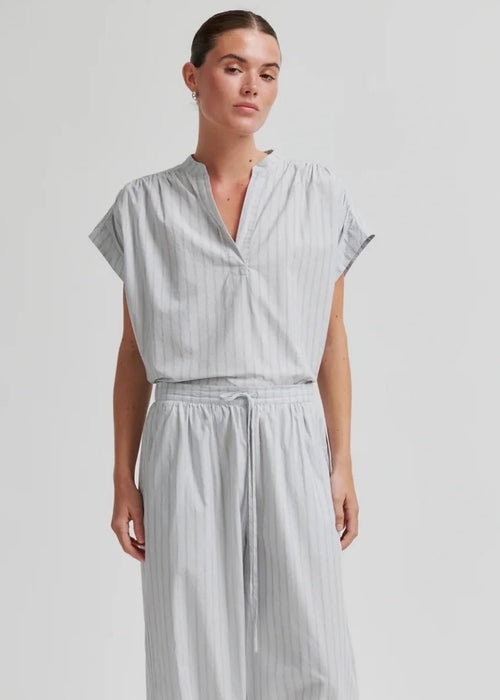 A woman modelling a light grey and white vertically striped two-piece outfit consisting of a short-sleeve V-neck top and matching loose harem-style pants, paired with black flip-flop sandals against a plain light grey studio background.