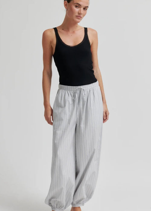 A woman modelling loose, light grey striped harem-style pants with a black tank top and black flip-flops against a plain light grey studio background.