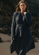Woman wearing a long dark blue cardi standing on a beach with rocks in the background