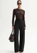 Woman wearing a brown long-sleeve top and black high-waisted pants, holding a black handbag on a white background.