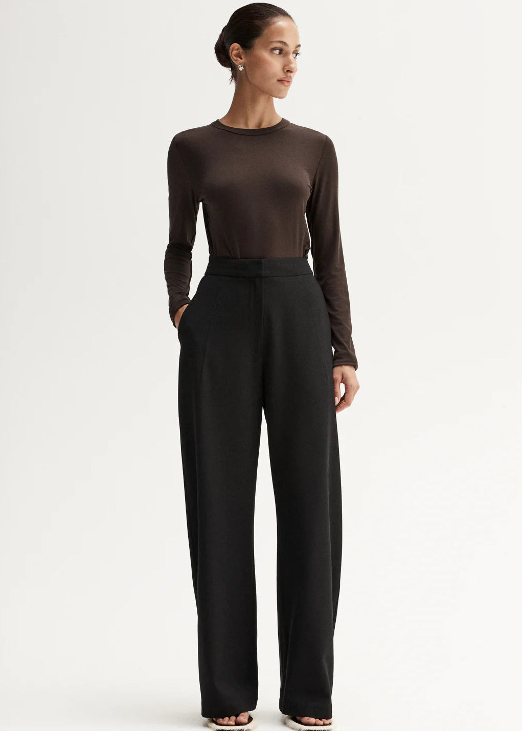 Woman wearing a brown long-sleeve top and black high-waisted pants, holding a black handbag on a white background.