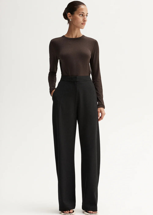 Woman wearing a brown long-sleeve top and black high-waisted pants, holding a black handbag on a white background.