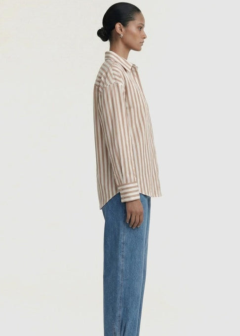 A model shown in profile wearing a beige and white vertically striped button-down shirt, blue wide-leg jeans, and brown sandals against a plain off-white studio background.