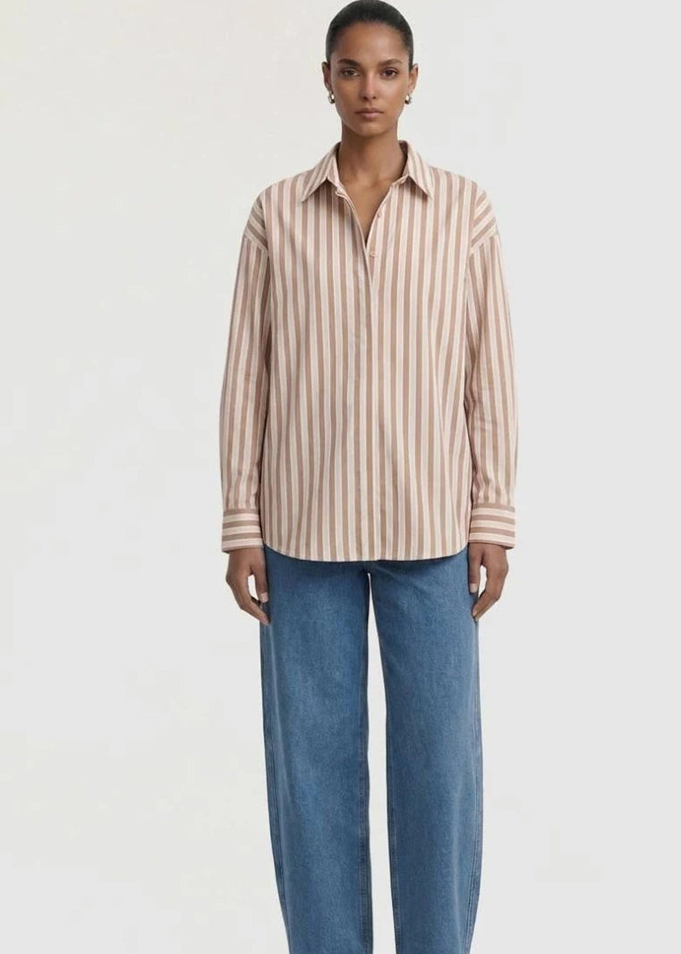 A model stands against a plain light background wearing a beige and white vertically striped button-down shirt, wide-leg blue jeans, and brown sandals.