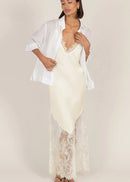 A woman modeling a cream lace slip dress with a sheer white button-up shirt draped over her shoulders, set against a solid light beige studio background.