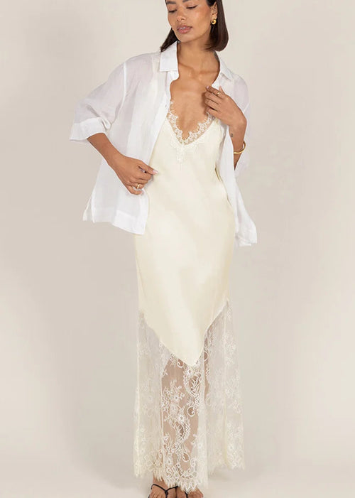 A woman modeling a cream lace slip dress with a sheer white button-up shirt draped over her shoulders, set against a solid light beige studio background.