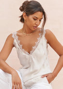 A woman wearing a white lace-trimmed camisole top and white pants, posed against a soft beige background.