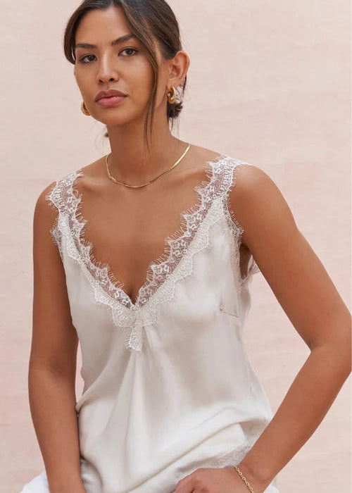 A woman wearing a white lace-trimmed camisole top and white pants, posed against a soft beige background.