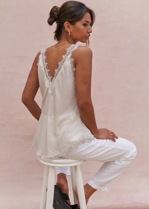 A woman wearing a white lace-trimmed camisole top and white pants, posed against a soft beige background.
