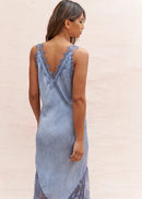 A woman wearing a light blue sleeveless slip dress with lace trim and a lace-up back, photographed from behind against a plain light beige studio background.