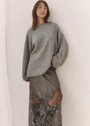 A woman stands against a white wall wearing a loose grey knit sweater and a long sheer grey skirt with black floral lace overlay and black pointed shoes.