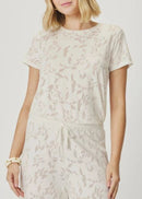 A woman wearing a matching short-sleeve t-shirt and drawstring pants set in a light cream color with a subtle beige botanical leaf pattern, photographed against a neutral light grey background.