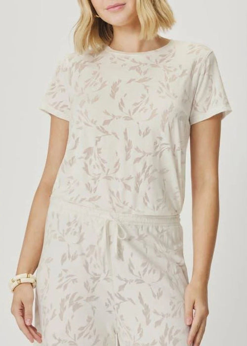 A woman wearing a matching short-sleeve t-shirt and drawstring pants set in a light cream color with a subtle beige botanical leaf pattern, photographed against a neutral light grey background.