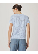 A woman viewed from the back wearing a light blue short-sleeve t-shirt with a subtle tonal leaf pattern and light-wash blue jeans, set against a plain light beige studio background.