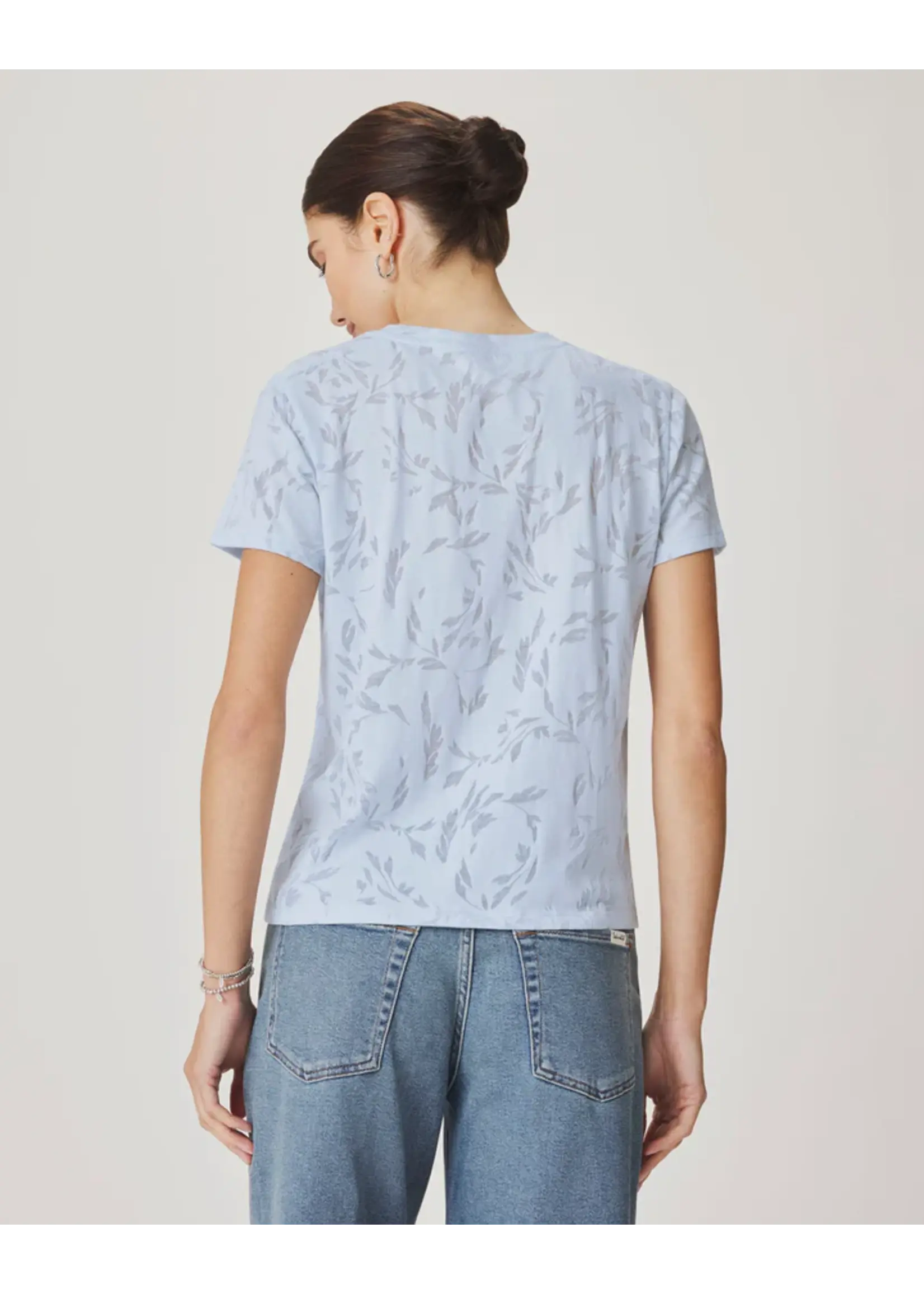 A woman viewed from the back wearing a light blue short-sleeve t-shirt with a subtle tonal leaf pattern and light-wash blue jeans, set against a plain light beige studio background.