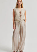 A woman modelling a sleeveless beige knit vest with front tie closures and matching wide-leg beige pants against a light grey studio background.