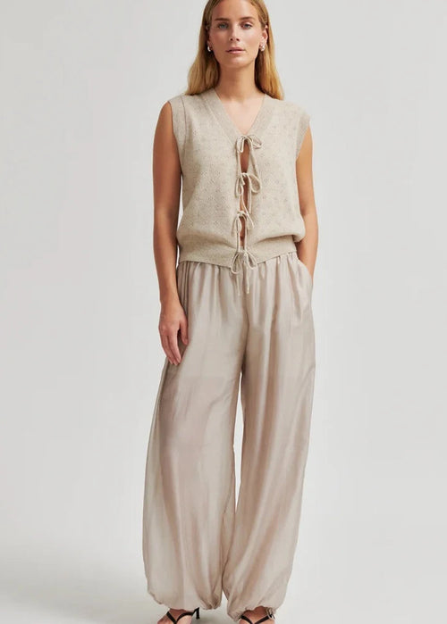 A woman modelling a sleeveless beige knit vest with front tie closures and matching wide-leg beige pants against a light grey studio background.