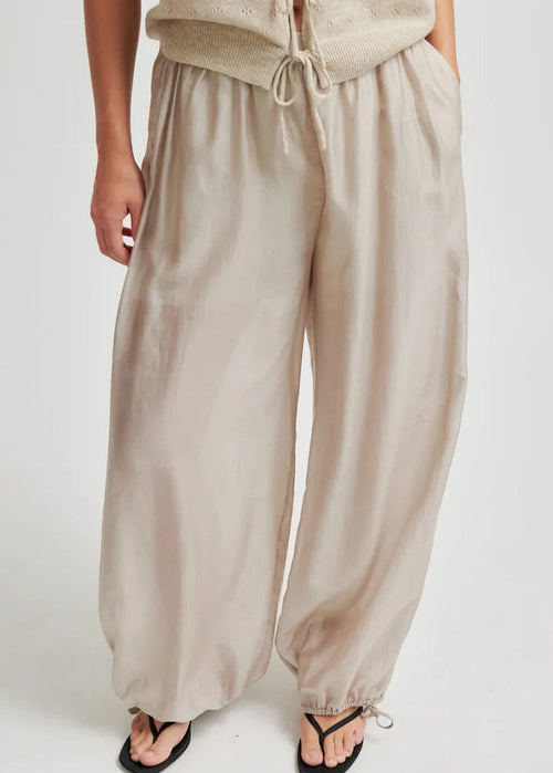 A person wearing loose, wide-leg beige pants with an elastic drawstring waist and gathered cuffs at the ankles, paired with black thong sandals against a plain light background.