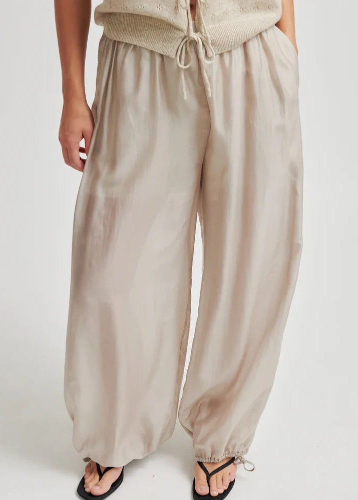 A person wearing loose, wide-leg beige pants with an elastic drawstring waist and gathered cuffs at the ankles, paired with black thong sandals against a plain light background.
