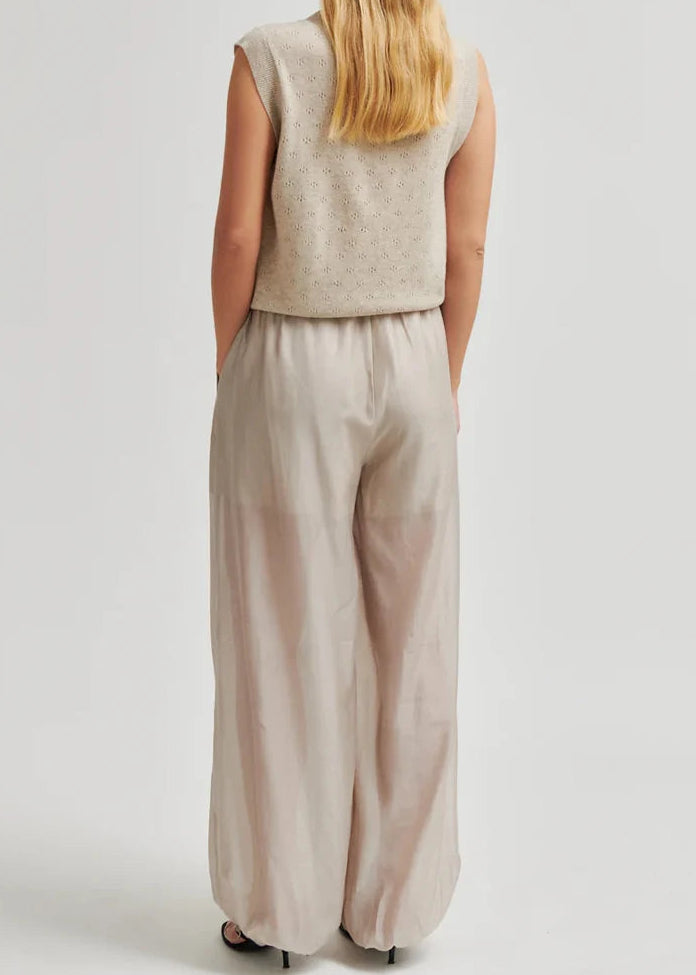 A woman modelling a sleeveless beige knit vest with front tie closures and matching wide-leg beige pants against a light grey studio background.