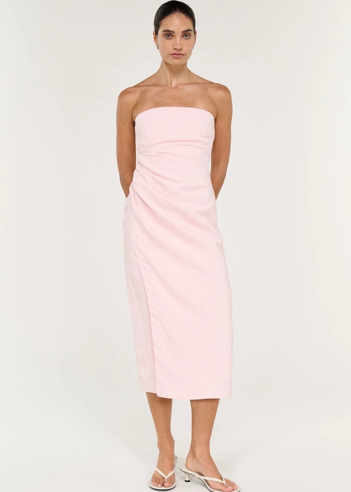 A woman modelling a pale pink strapless midi dress with gathered draping at the waist, photographed against a clean off-white studio background.