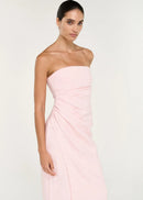 A woman modelling a pale pink strapless midi dress with gathered draping at the waist, photographed against a clean off-white studio background.