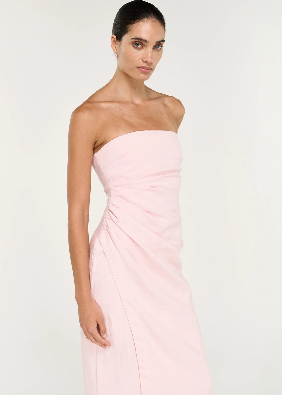 A woman modelling a pale pink strapless midi dress with gathered draping at the waist, photographed against a clean off-white studio background.