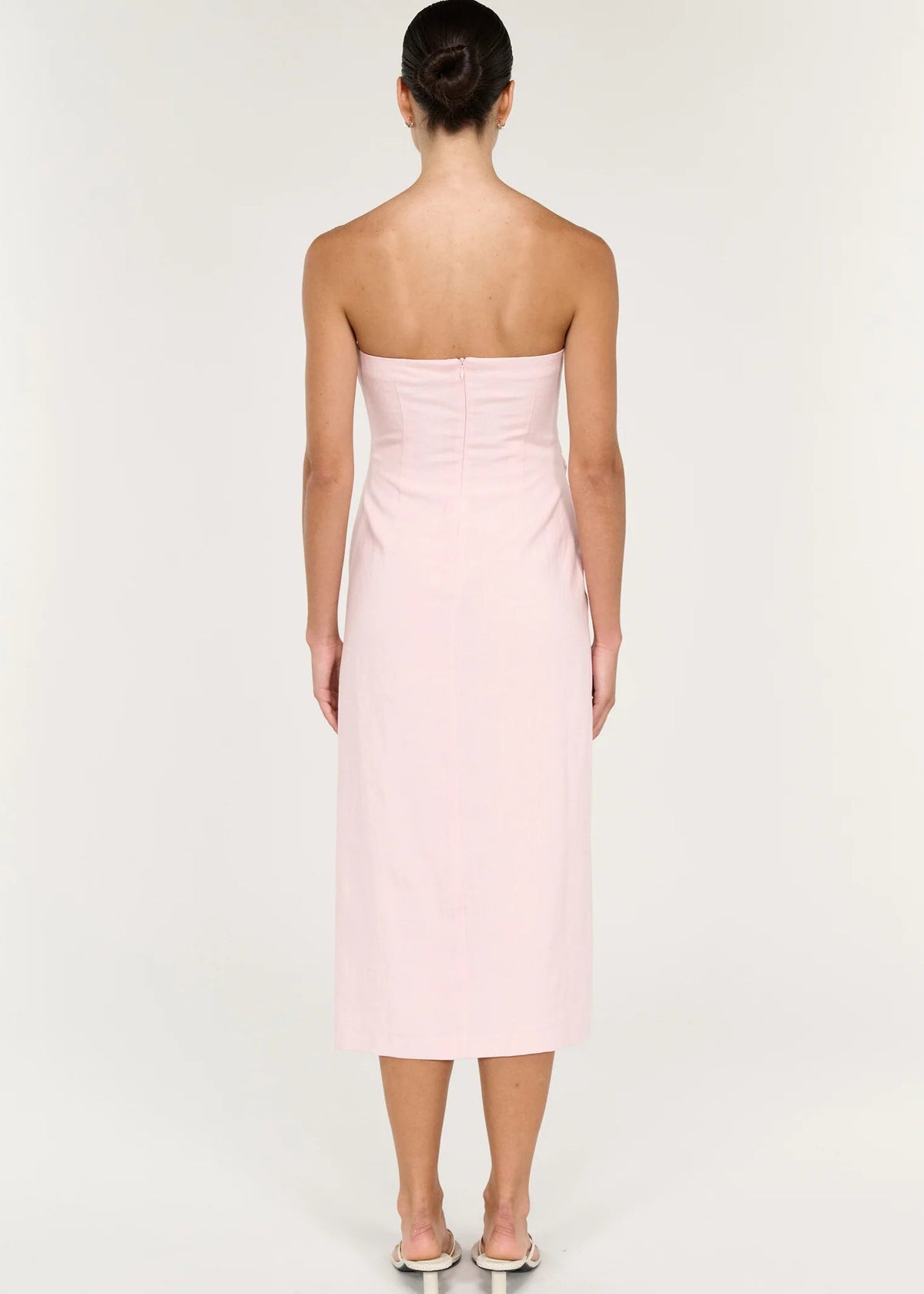 A woman modelling a pale pink strapless midi dress with gathered draping at the waist, photographed against a clean off-white studio background.