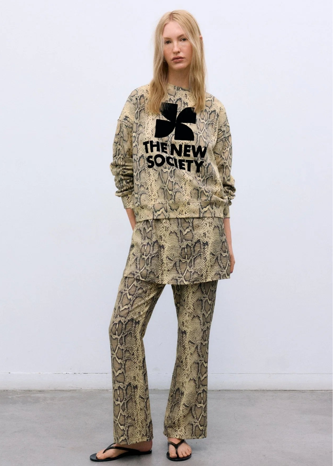 A model stands against a plain white wall wearing a matching two-piece outfit with a beige and black snakeskin pattern. The set includes a long-sleeve sweatshirt with a black four-leaf clover graphic and the text 'THE NEW SOCIETY' across the chest, paired with matching wide-leg pants.