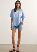 A woman wearing a light blue oversized graphic t-shirt with the French phrase 'TOUT VA BIEN' printed in white block letters with a small red heart accent, paired with blue denim shorts.